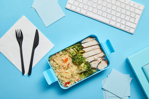 Top view of lunch box with delicious rice with chicken and broccoli at workplace with computer keyboard on blue background