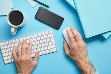 Mavi arka plan üzerinde kağıtları, akıllı telefon ve kahve fincan ı ile işyerinde bilgisayar klavyesi ve bilgisayar faresi kullanan adamın kırpılmış görünümü