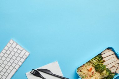 Mavi arka plan üzerinde risotto, tavuk ve brokoli ile bilgisayar klavyesi ve öğle yemeği kutusu ile işyeri üst görünümü