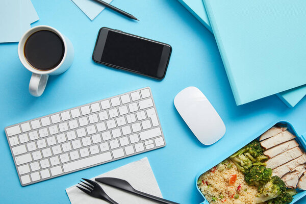 Top view of workplace with digital devices, papers and lunch box with rice, broccoli and chicken on blue background