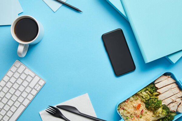 Top view of workplace with digital devices, papers and lunch box with healthy and tasty food on blue background