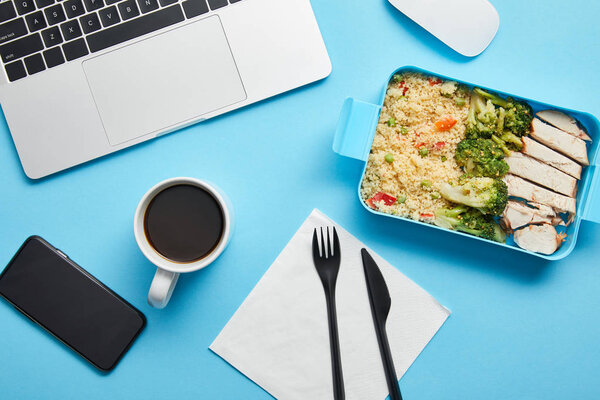 Top view of workplace with digital devices, cup of coffee and lunch box with rice, broccoli and chicken on blue background,illustrative editorial
