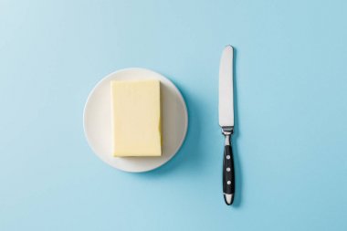 top view of butter on white plate and knife on blue background clipart