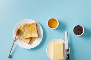 top view of butter, knife on plate, jam, bowls, toasts with honey and wooden dipper on white plates on blue background clipart