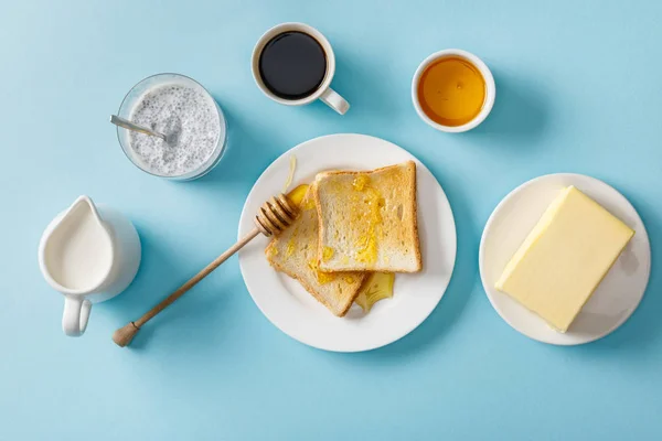 コーヒー ミルク チアシード バター 青い背景に白いプレートに蜂蜜と木製のディッパーとトーストのトップビュー — ストック写真