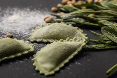 selective focus of green ravioli near sage, flour and pine nuts on black table clipart