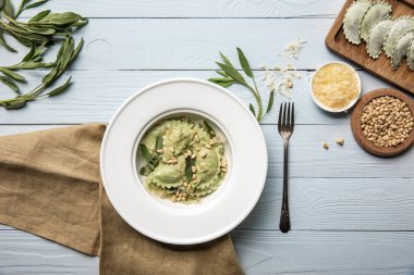 çatal ve çuval bezi ile beyaz ahşap masa üzerinde çam fıstığı, adaçayı ve rendelenmiş peynir ile beyaz tabakta servis yeşil ravioli üst görünümü