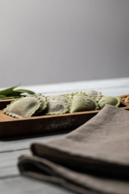 selective focus of raw green ravioli served on wooden board near napkin isolated on grey clipart