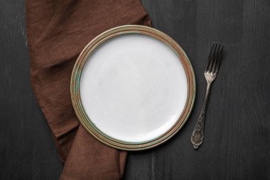 kahverengi peçete ve gümüş çatal yakınında siyah ahşap masa üzerinde vintage beyaz boş yuvarlak plaka üst görünümü