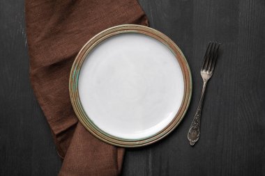 top view of vintage white empty round plate on black wooden table near brown napkin and silver fork clipart