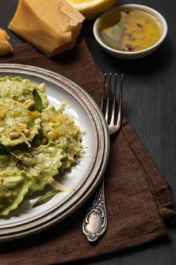 close up view of delicious green ravioli with sage, cheese and pine nuts served on black wooden table with fork and napkin clipart