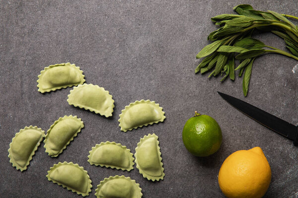 top view of green ravioli near lemon, lime, knife and herb on grey textured surface