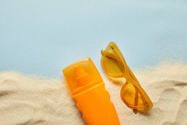 top view of sunscreen in orange bottle near sunglasses on blue background with sand clipart