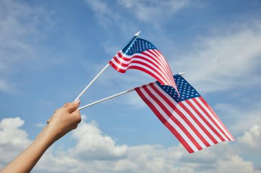 Cropped view of  woman holding american flags in hand on blue sky background clipart