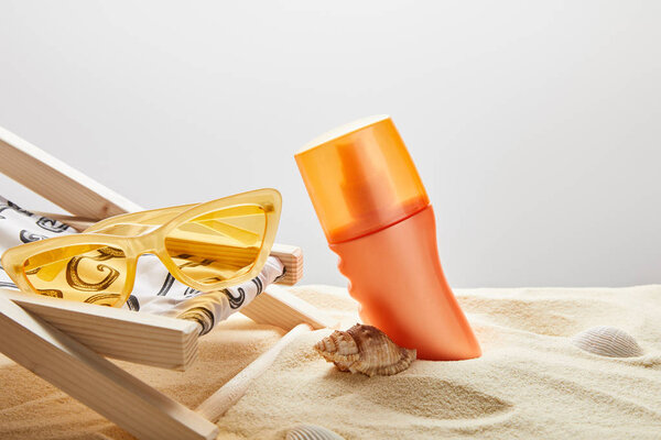 оранжевый солнцезащитный крем в песке рядом с раковинами, желтые солнцезащитные очки и шезлонг на сером фоне
