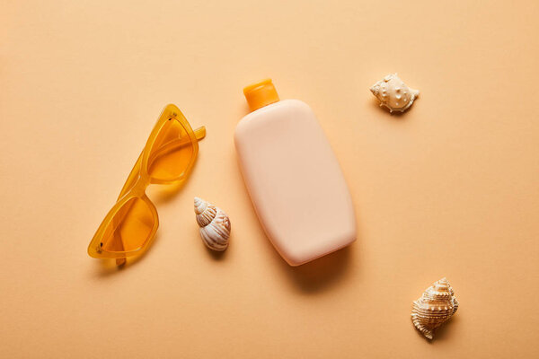 top view of sunscreen lotion in bottle near scattered seashells and sunglasses on beige background