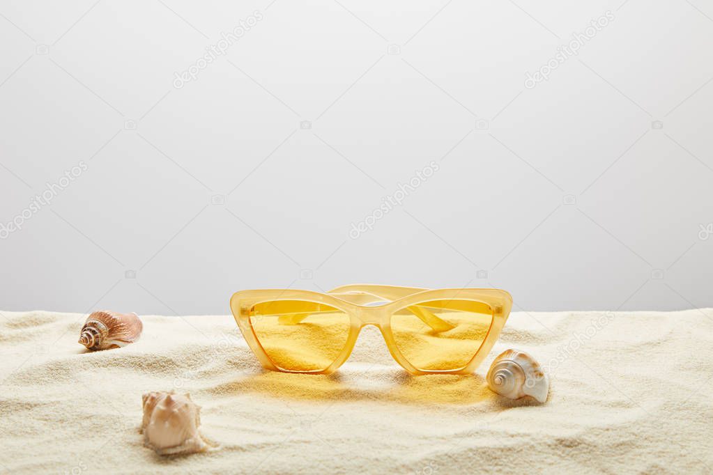 yellow stylish sunglasses on sand with seashells on grey background