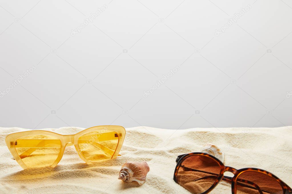 yellow and brown stylish sunglasses on sand with seashells on grey background