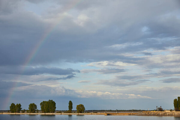 landscape of blue sunlight sky, rainbow, river and coast with trees