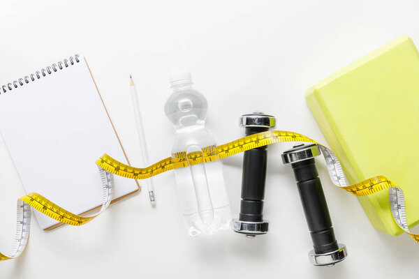 top view of water in bottle, measuring tape, dumbbells and blank notebook with pencil near box on white background 