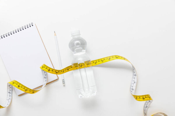 top view of water in bottle, measuring tape, blank notebook with pencil on white background 