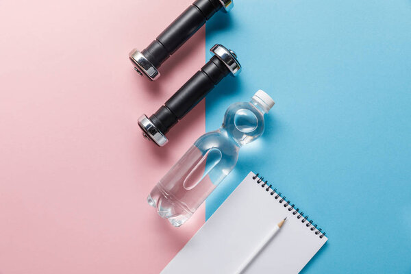 top view of bottle with water, dumbbells and blank notebook on blue and pink background