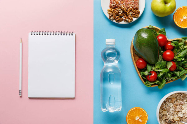 top view of bottle with water and diet food near blank notebook on blue and pink background