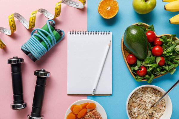 top view of fresh diet food near sport equipment and measuring tape around blank notebook on pink and blue background