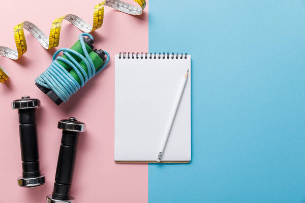 top view of notebook, dumbbells, skipping rope and measuring tape on blue and pink background 