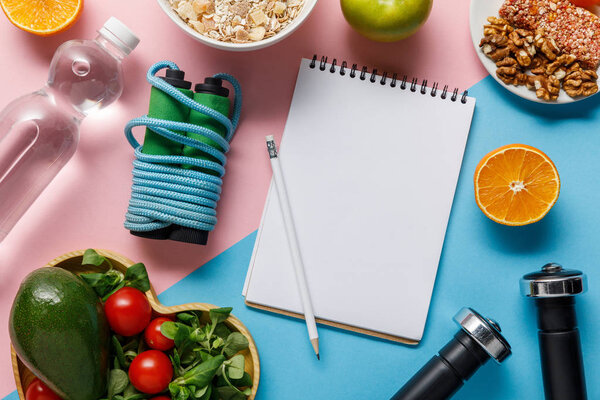 вид на пустой блокнот и карандаш с вкусной диетической пищей, водой и спортивным оборудованием на синем и розовом фоне
 