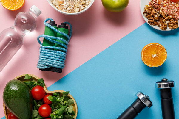 top view of diet food, water, skipping rope and dumbbells on pink and blue background