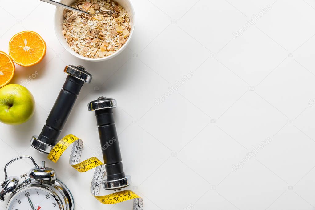 top view of dumbbells, measuring tape, alarm clock and breakfast cereal with fruits on white background