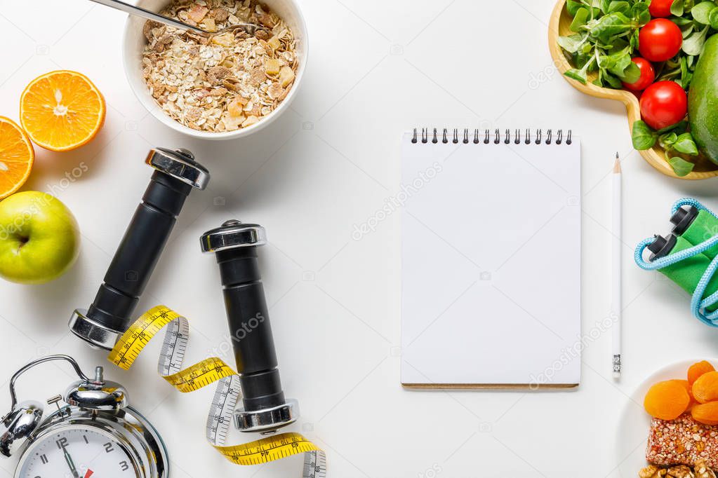 top view of sport equipment, measuring tape, alarm clock and diet food near blank notebook on white background