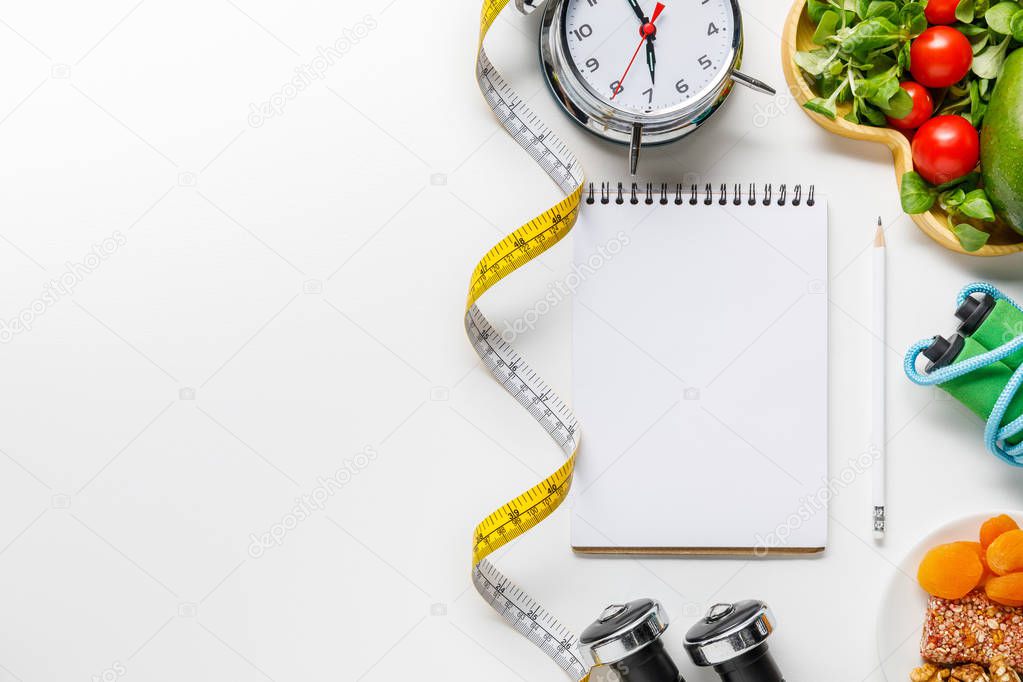 top view of sport equipment, measuring tape, alarm clock and diet food near empty notebook on white background