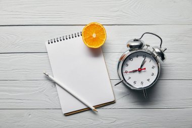 top view of orange half, blank notebook and pencil near alarm clock on wooden white background clipart