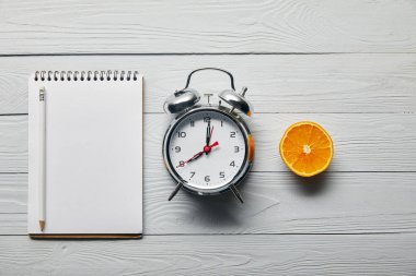 gümüş çalar saat, ahşap beyaz arka plan üzerinde kalem ve turuncu yarısı ile boş defter ile düz yatıyordu 