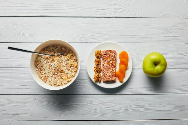 平らなボウルに朝食シリアル ナッツと木製の白い背景に緑のリンゴの近くに乾燥したアプリコットとプレート — ストック写真