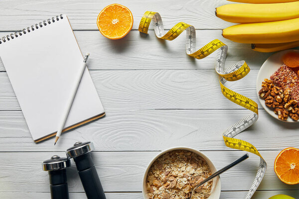 top view of measuring tape, dumbbells, diet food and blank notebook on wooden white background 