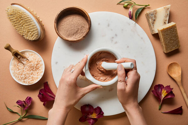 cropped view of woman mixing clay in bowl on marble circle near organic cosmetic ingredients on beige background with scattered flower petals