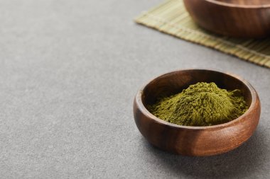 selective focus of green matcha powder in wooden bowl on grey table with copy space clipart