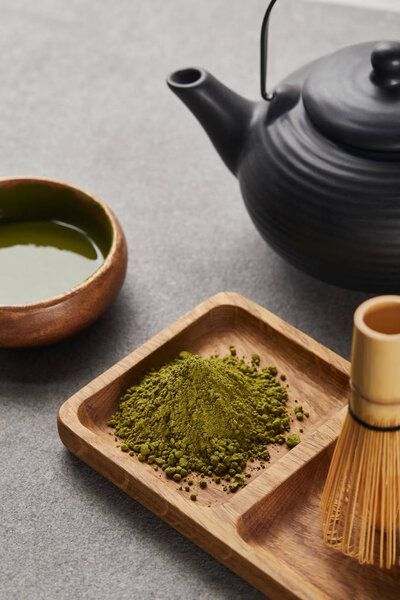 selective focus of green matcha powder and bamboo whisk on wooden board near black teapot and bowl with tea