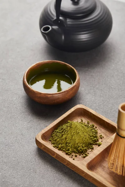 green matcha powder and bamboo whisk on wooden board near black teapot and bowl with tea