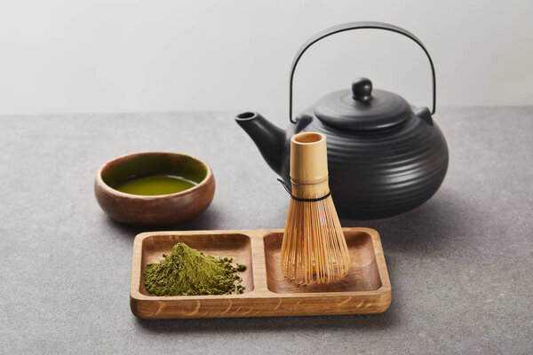 green matcha powder and bamboo whisk on wooden board near black teapot and bowl with tea