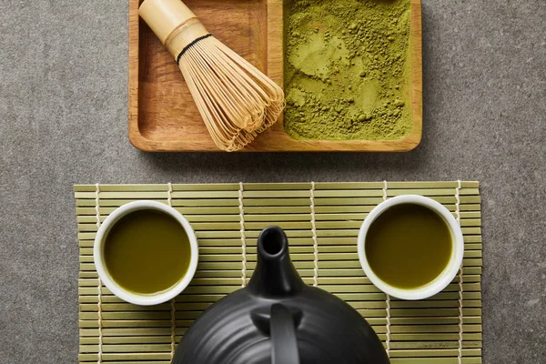 top view of white cups with tea and black teapot on green table mat near bamboo whisk and matcha powder on board