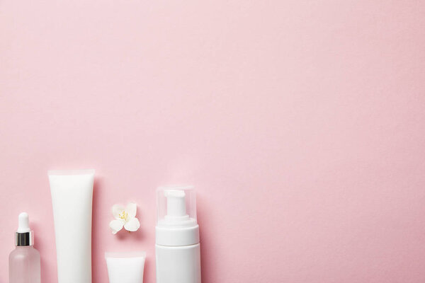 вид сверху на пустую бутылку из-под косметического стекла, сливочные трубки со сливками, дозатор для косметики и цветок жасмина на розовом
 