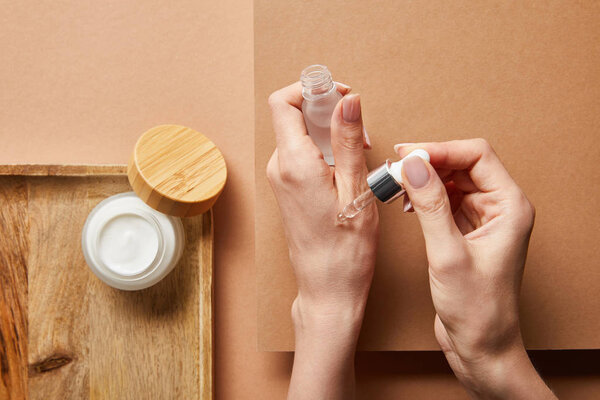 cropped view of woman holding cosmetic glass bottle with serum near open jar with cream on wooden tray on brown 