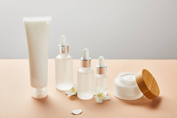 cream tube, cosmetic glass bottles with serum and open jar with cream near few jasmine flowers