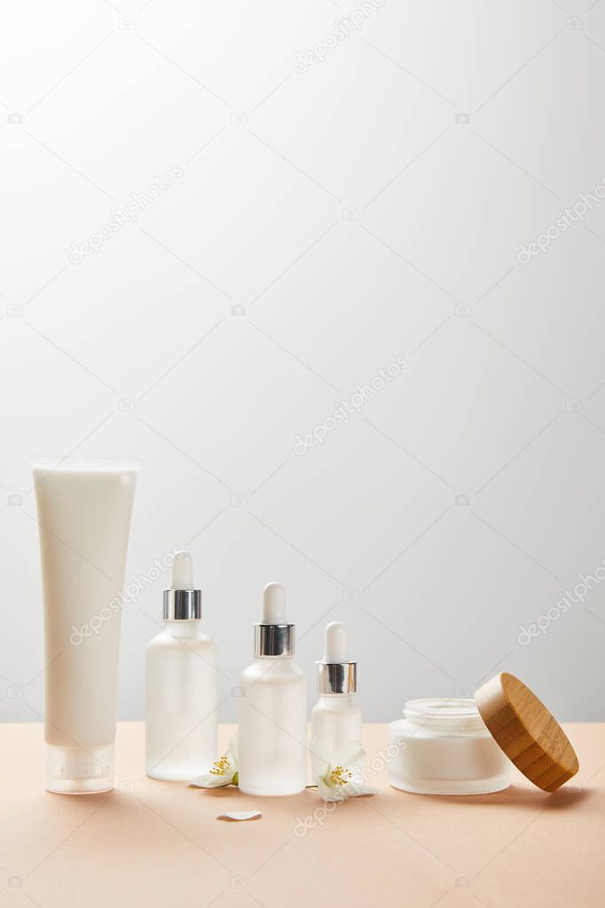 cream tube with cream, cosmetic glass bottles with serum, open jar with wooden cap and few jasmine flowers on beige 