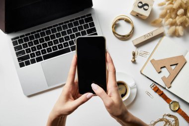 bitkiler, dizüstü bilgisayar, not defteri, saat, kahve ve kırtasiye ile beyaz yüzeyüzerinde akıllı telefon ile çalışan kadın kırpılmış görünümü