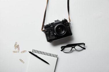 top view of photo camera, glasses, notepad with pen and stationery on white surface clipart